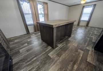 Spacious room with wood-look flooring, a large central kitchen island with a rustic finish, and three windows with brown and white curtains letting in natural light. Ceiling fan visible in the background.