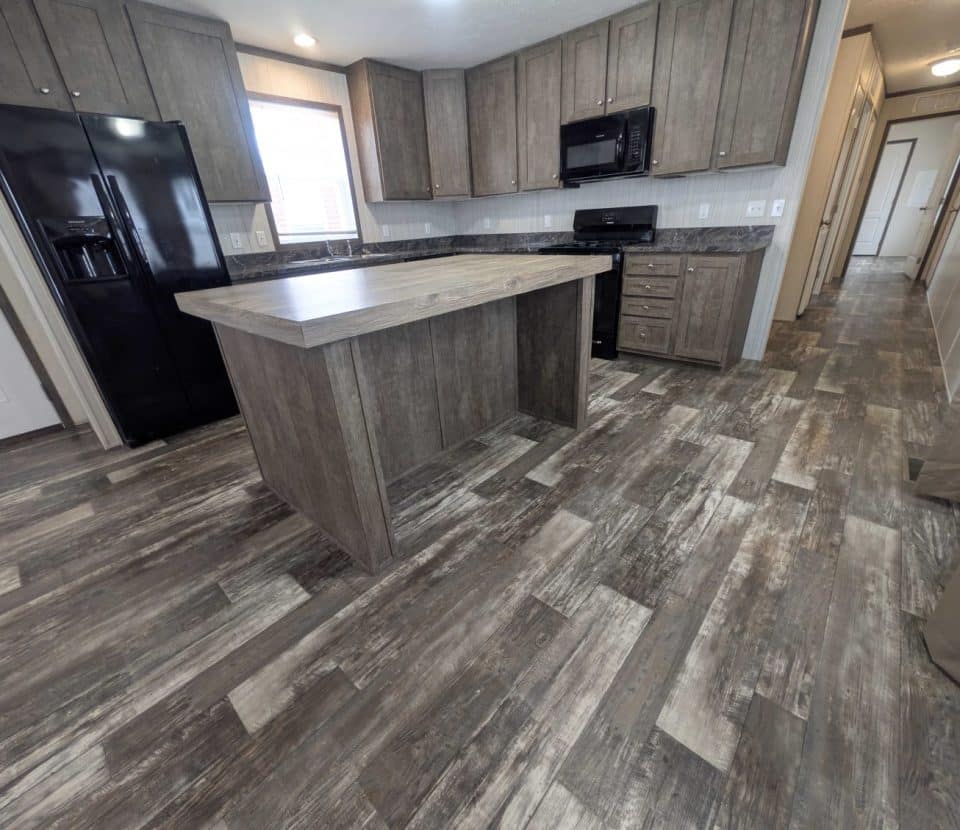 Spacious modern kitchen with wood-look tile flooring, gray cabinets, a center island, black appliances, and ample natural light coming through a window above the sink. Hallway and multiple doors visible in the background.