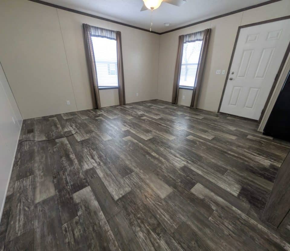 Empty room with wood-look vinyl flooring, cream-colored walls, two windows with brown curtains, a white door, and a ceiling fan; partial view of a kitchen area with black appliances on the right.