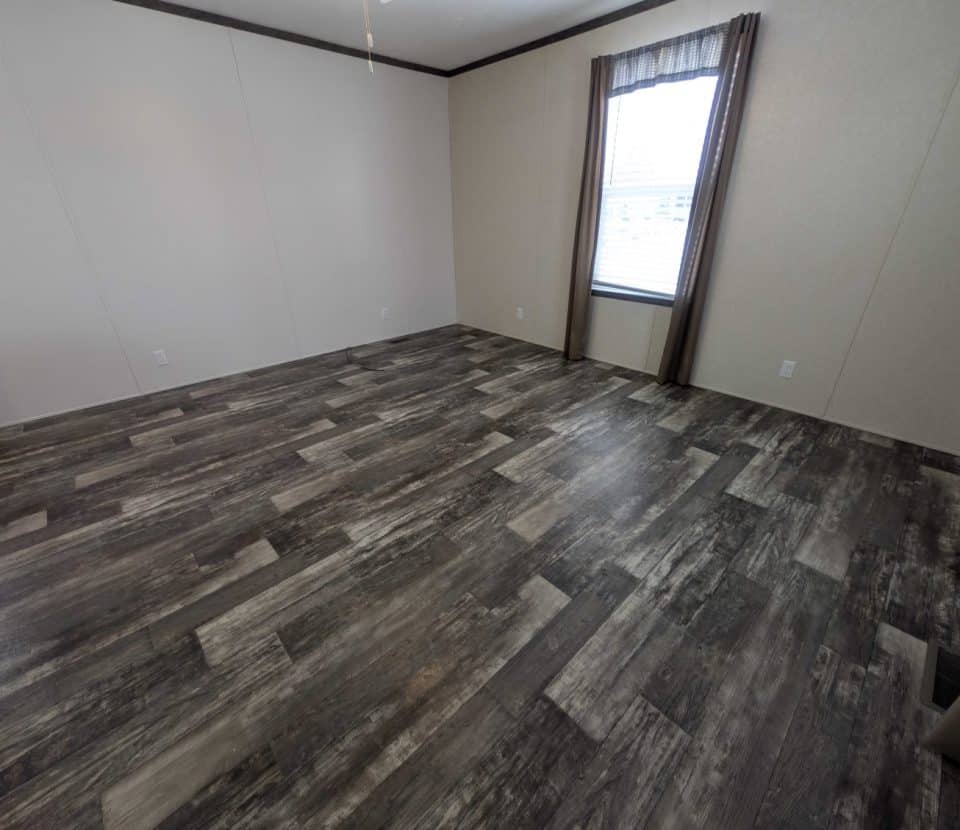 Empty room with light-colored walls, a large window with sheer curtains, and dark wood-patterned vinyl flooring. The space is well-lit with natural light coming through the window.