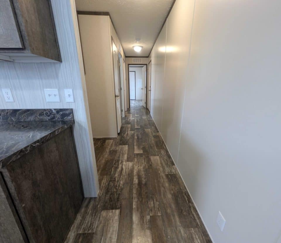 Long hallway with brown wood-style flooring, beige walls, doors leading to rooms, and a dark marble countertop with cabinets on the left side. Ceiling light is visible at the far end.