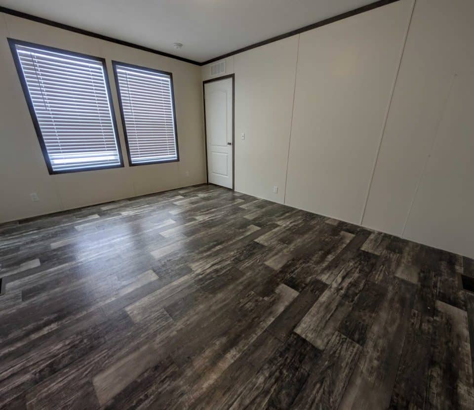 An empty room with dark wood-patterned flooring, cream-colored walls, two large windows with closed blinds, and a white door. The room is well-lit and unfurnished.
