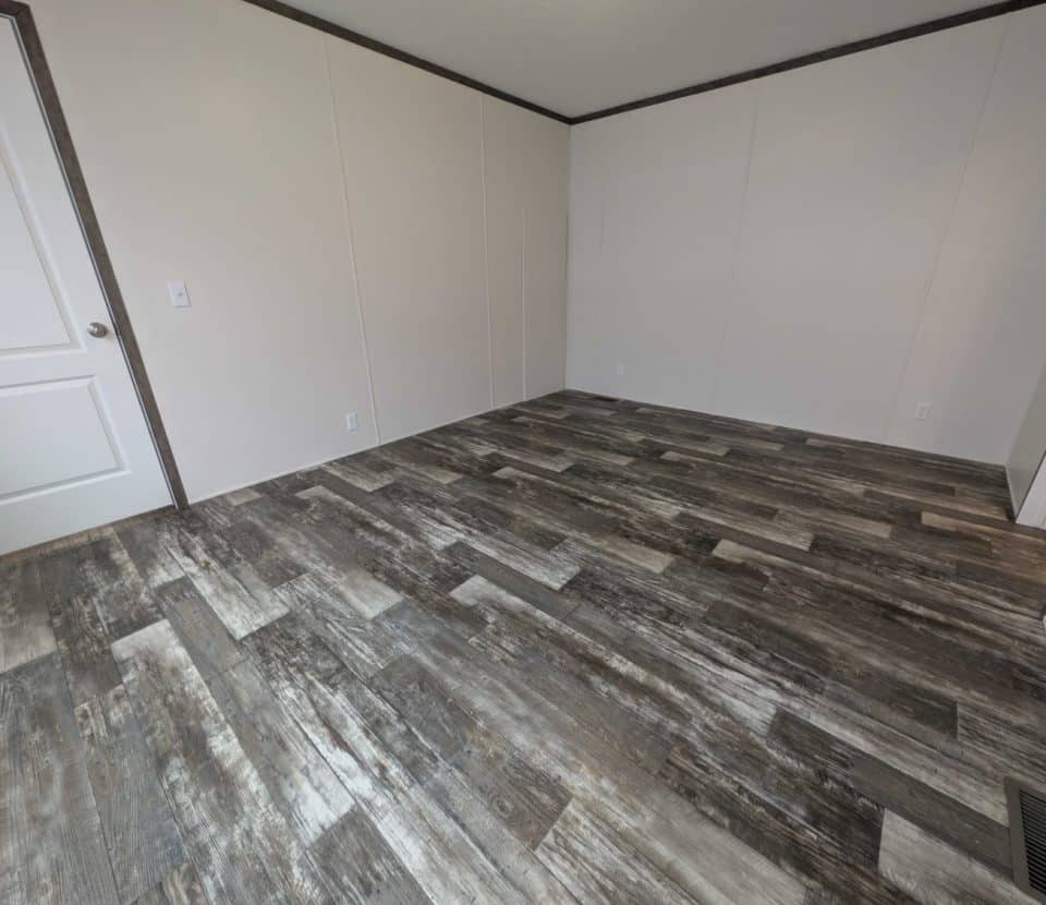 An empty room with light-colored walls, dark trim, and rustic wood-look vinyl flooring. The room has a closed white door, electrical outlets, and an air vent on the floor.