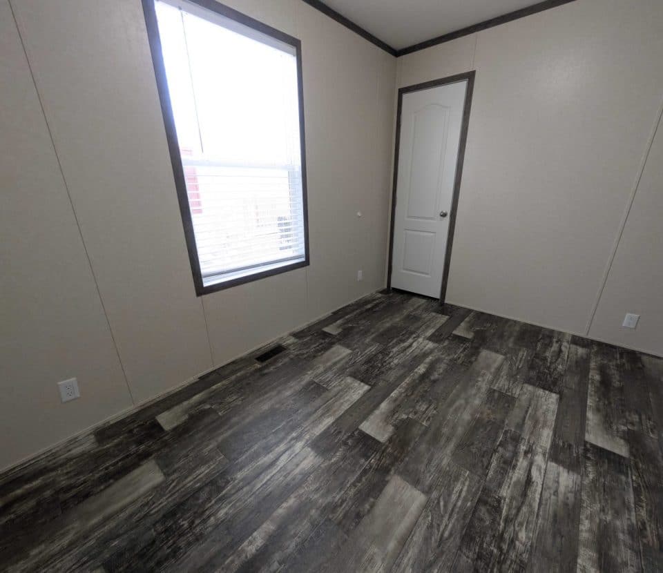 Empty room with beige walls, a white door, a large window with blinds, and dark wood-patterned vinyl flooring. Electrical outlets are visible on the walls, and natural light enters through the window.