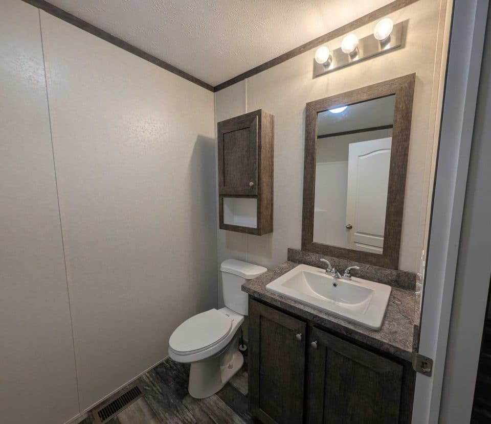 A bathroom with a toilet, a sink with a dark wood vanity, a matching mirror, and a wall cabinet. The lighting fixture above the mirror has three bulbs, and the room has grayish-brown flooring and light-colored walls.