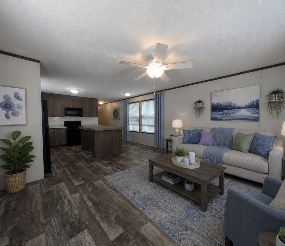 A modern open-plan living room and kitchen with wood flooring, neutral walls, a ceiling fan, a beige sofa with colorful pillows, a blue armchair, a coffee table, and kitchen appliances in the background.