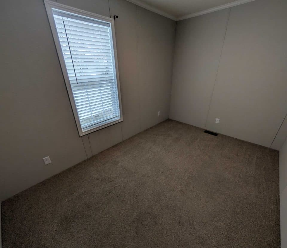 An empty room with beige carpet, light gray walls, a single window with white blinds, and several electrical outlets. The room appears clean and unfurnished.