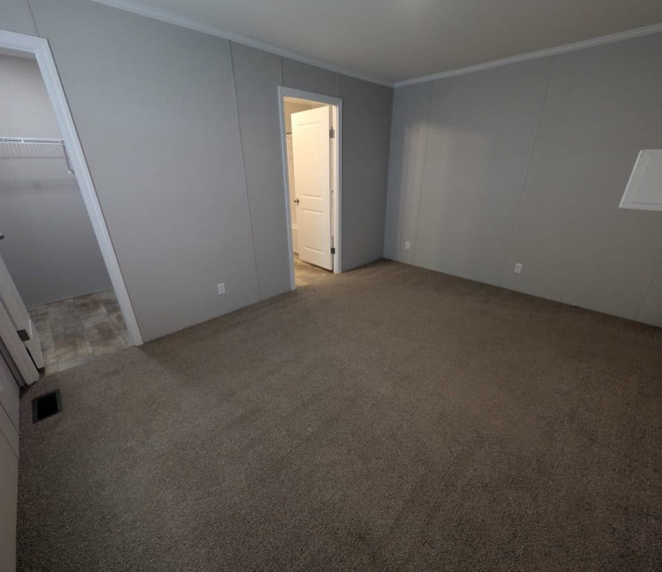 Empty carpeted room with light gray walls, an open door leading to a walk-in closet, another door leading to a bathroom, and a white panel on one wall. Ceiling has white trim. Room is well-lit.
