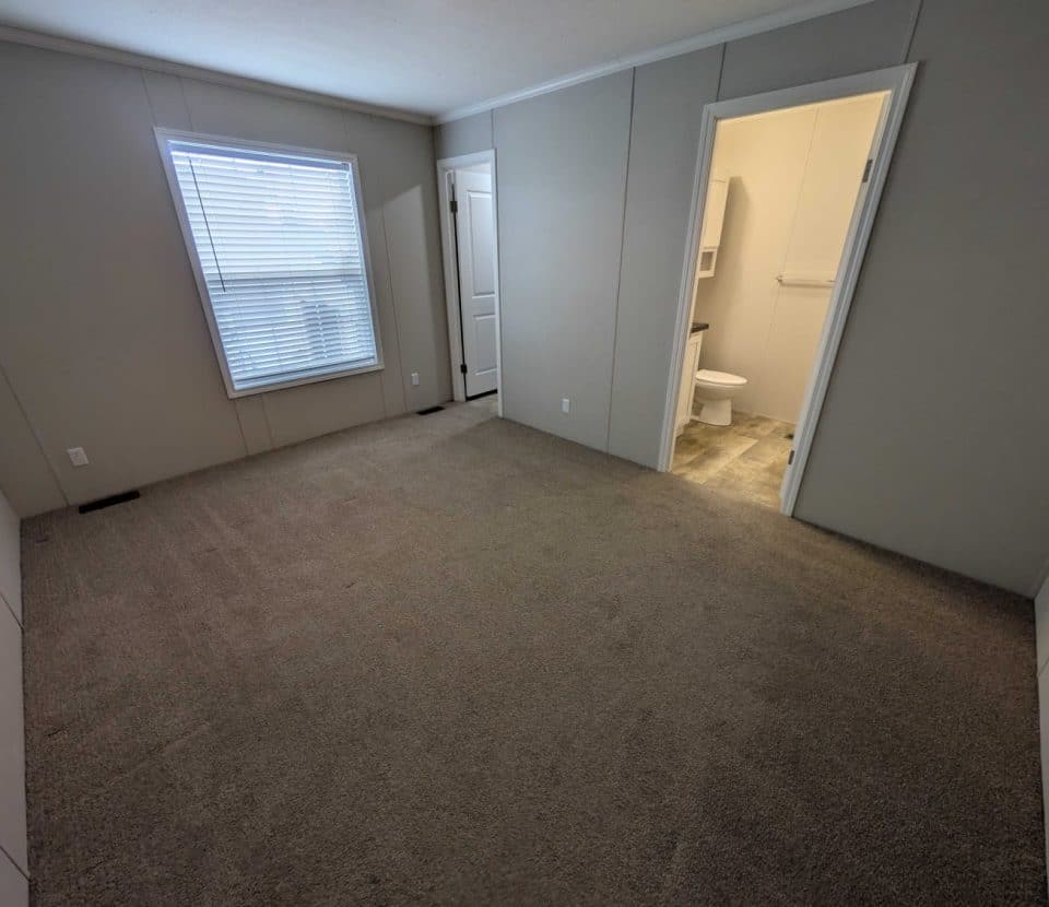 A carpeted, empty bedroom with a window on the left wall; a door leads to a small bathroom with a toilet and shelves. The walls are light-colored, and the room is well-lit by natural light.