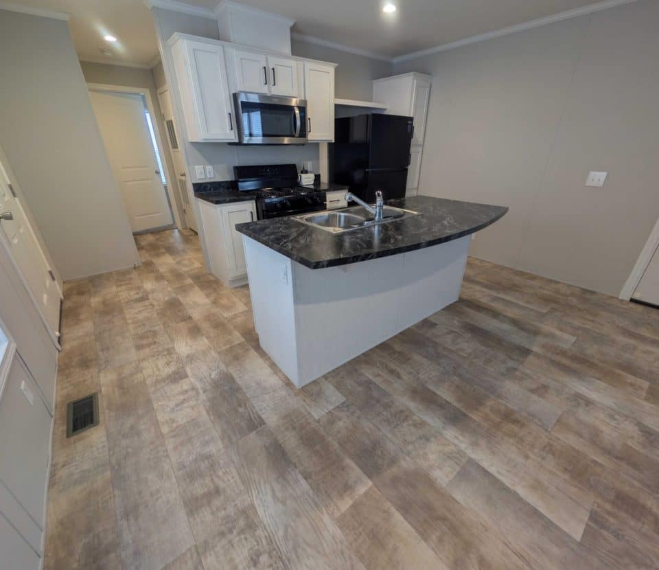 Modern kitchen with light wood cabinets, black appliances, a marble-look island with a sink, and wood-patterned vinyl flooring; doorways and bright overhead lighting are visible.