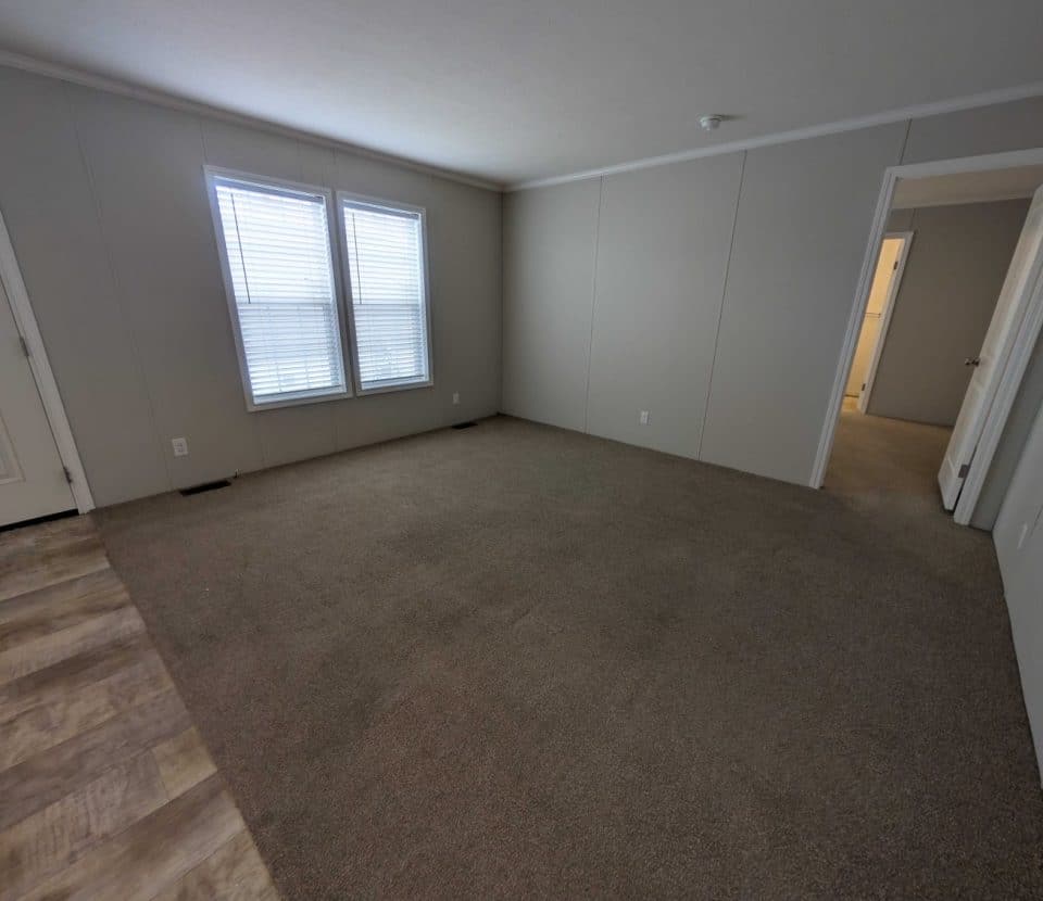 Empty room with beige carpet and light-colored walls, two windows with blinds letting in natural light, a white door, and a section of vinyl flooring leading to a hallway with open doors.