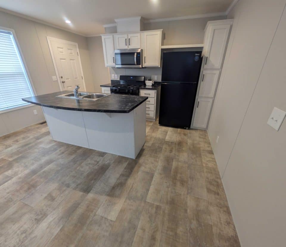 Modern kitchen with wood-look flooring, an island with a sink, white cabinets, black refrigerator, stove, and microwave. Large window on the left lets in natural light; neutral-colored walls.