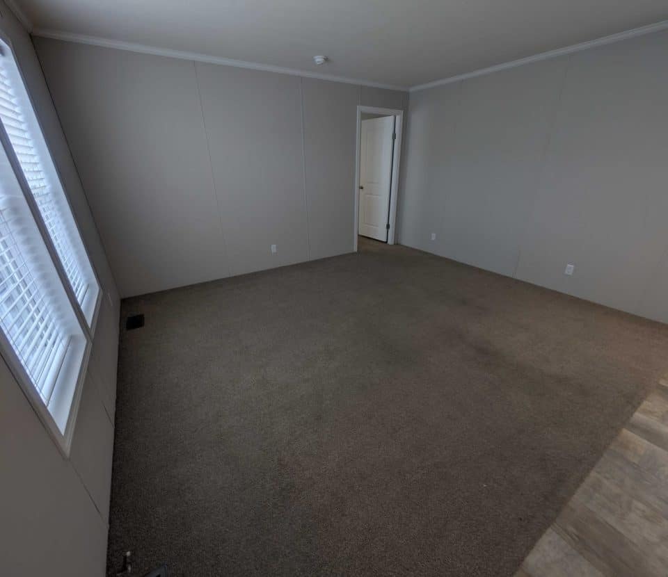 Empty room with beige carpet, light gray walls, large window with blinds on the left, white ceiling, and a door leading to another area. Part of the floor near the right edge transitions to a wood pattern.