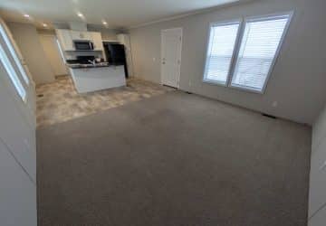 Spacious open-concept room with carpeted living area and tiled kitchen. The kitchen features white cabinets, stainless steel appliances, and an island. Large windows and a door are on one wall, letting in natural light.