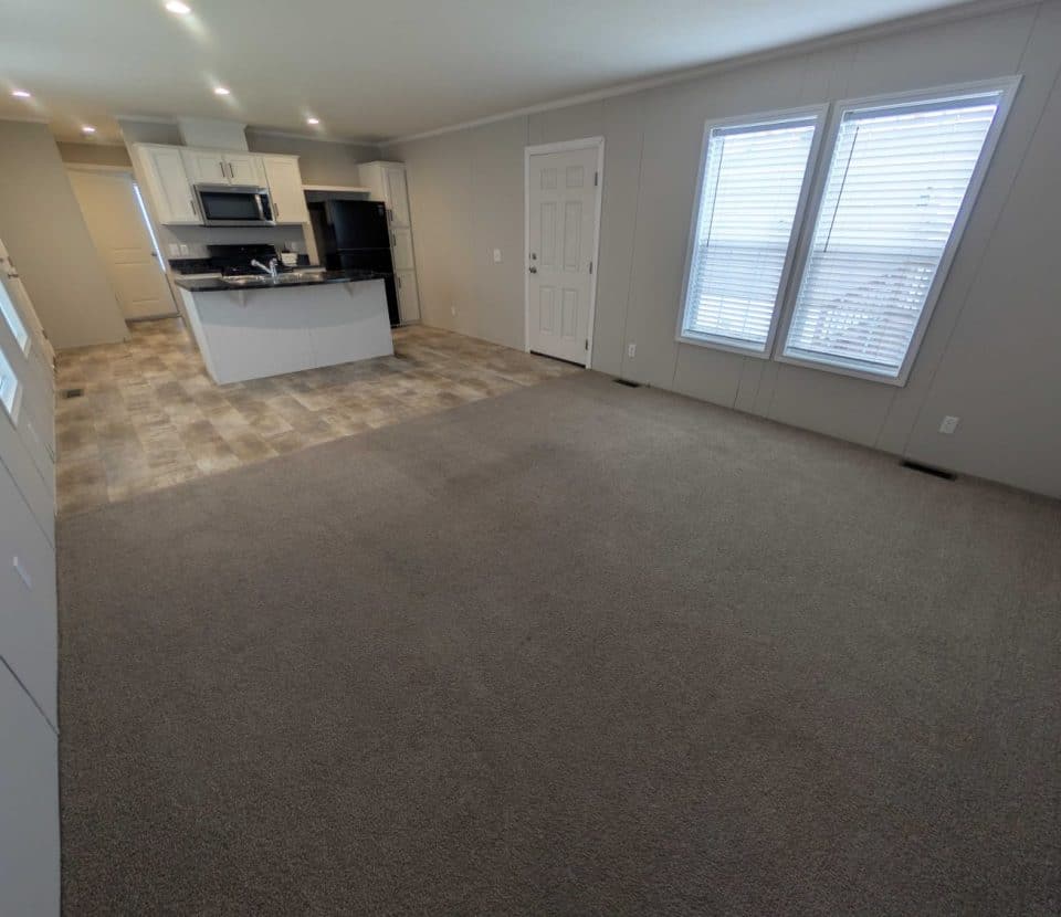 Spacious open-concept room with carpeted living area and tiled kitchen. The kitchen features white cabinets, stainless steel appliances, and an island. Large windows and a door are on one wall, letting in natural light.