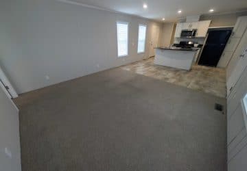 Open living area with brown carpet leading into a kitchen with tile flooring, white cabinets, stainless steel appliances, and a black refrigerator. Two tall windows and a white door are visible in the background.