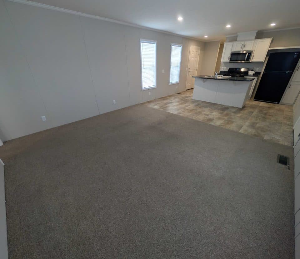 Open living area with brown carpet leading into a kitchen with tile flooring, white cabinets, stainless steel appliances, and a black refrigerator. Two tall windows and a white door are visible in the background.