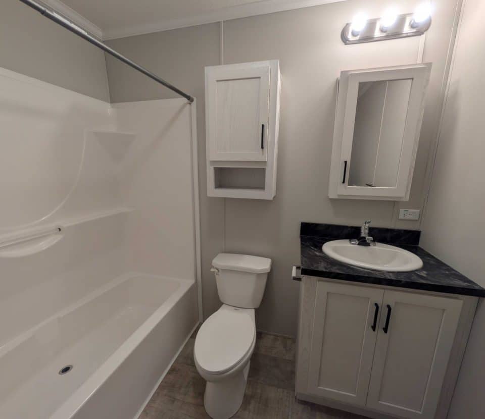 A modern bathroom with a white bathtub, a toilet, a vanity with a sink, a mirrored medicine cabinet, and overhead lighting. The walls and cabinets are light-colored, and the countertop is dark.
