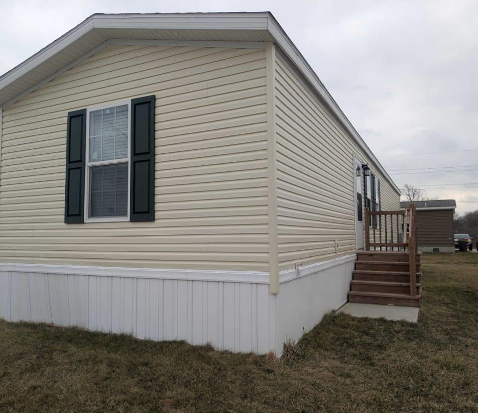 A beige manufactured home with white trim, green shutters, and a small wooden staircase leading to the entrance, sits on a grassy lot under a cloudy sky.