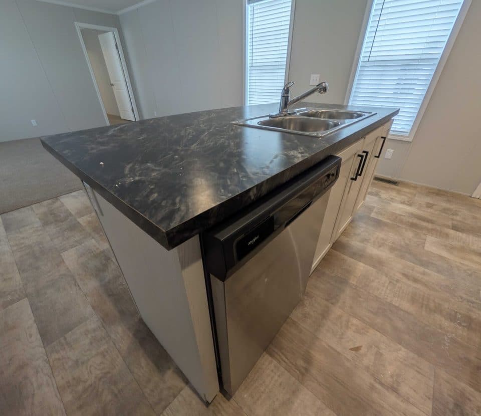 A kitchen island with a dark marble countertop, double sink, and stainless steel dishwasher, situated on wood-patterned flooring with windows in the background.