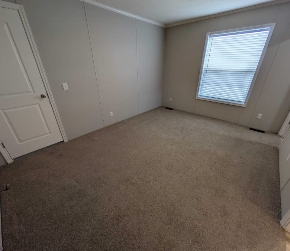 An empty room with beige carpet, light-colored walls, a closed white door, and a window with closed blinds letting in some daylight. The room appears clean and unfurnished.