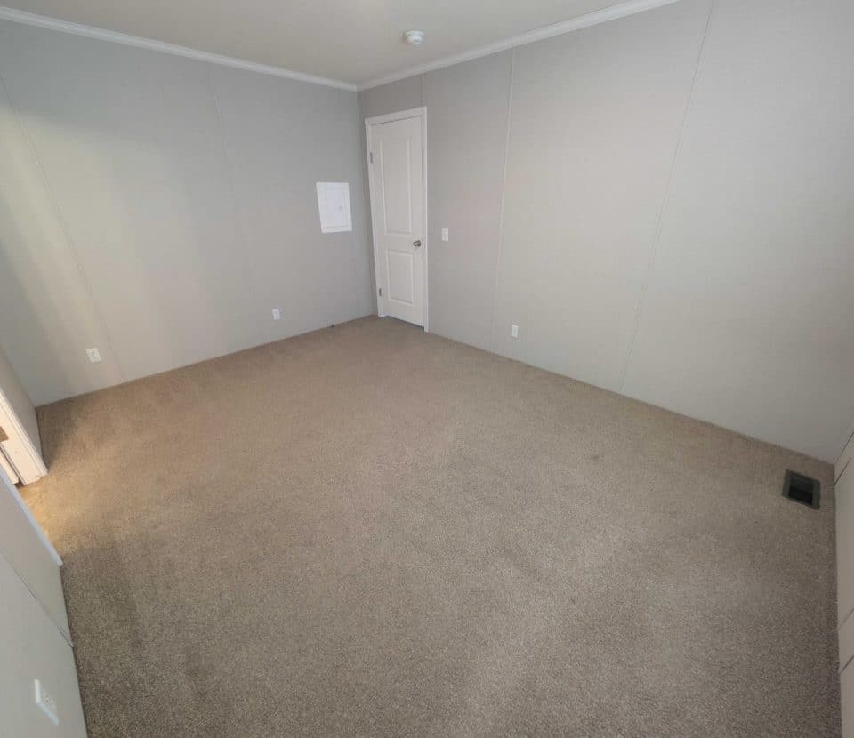 Empty room with beige carpet, light gray walls, a white door, and a small vent on the floor. The room has no furniture and is well-lit with natural light from the left side.
