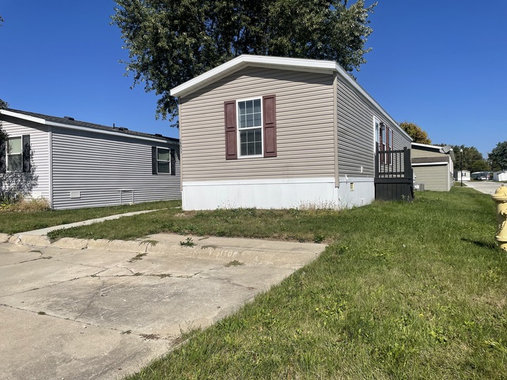 A beige mobile home with maroon shutters sits on a grassy lot beside a concrete driveway under a bright blue sky. Another mobile home and a yellow fire hydrant are visible nearby.