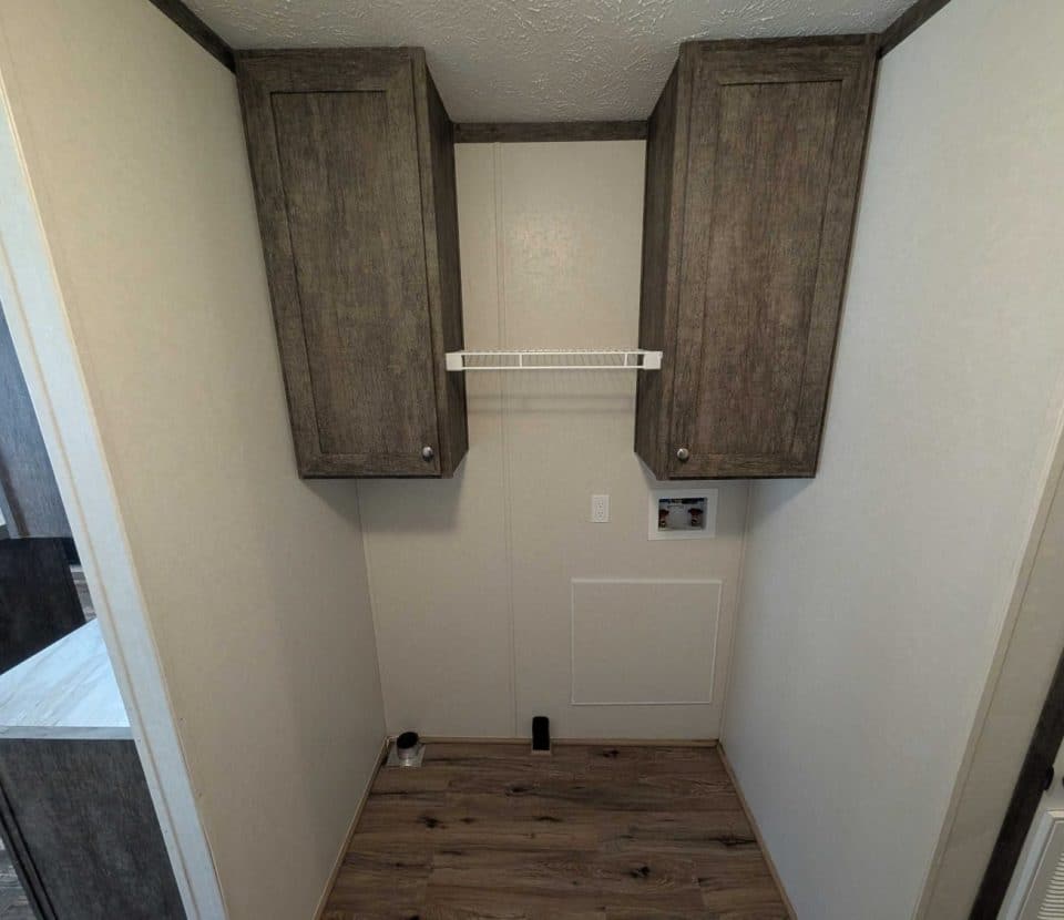 A small laundry room with wooden floor, two tall brown cabinets on the wall, a white wire shelf in between, and utility hook-ups on the wall below. The space is empty and has light-colored walls.