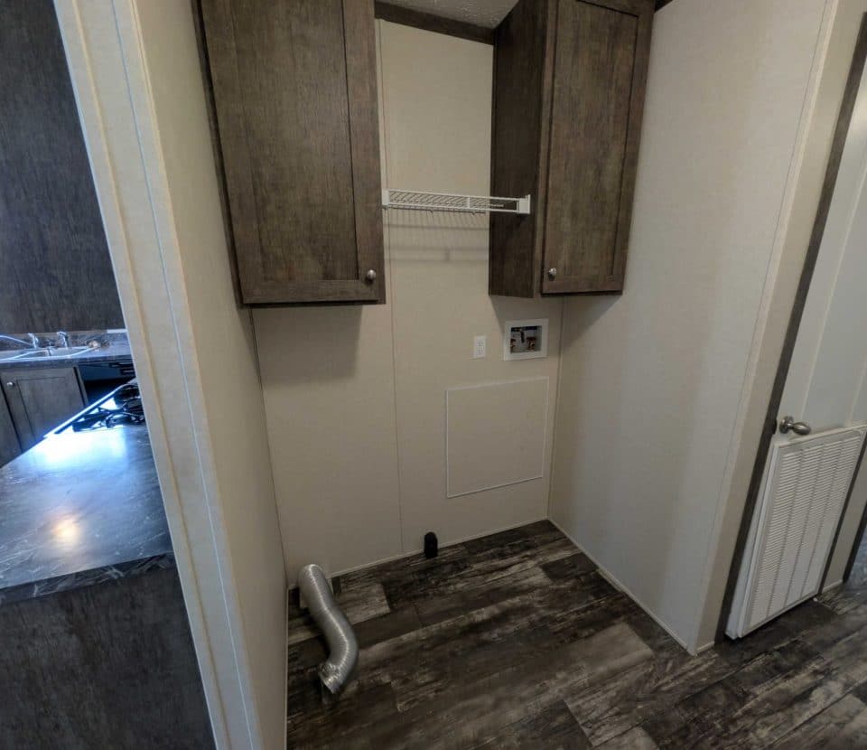 Laundry area with two wooden cabinets, a small wire shelf, washer and dryer hookups, and an exposed vent on a wood-patterned floor. Kitchen is visible to the left, and a door to the right.