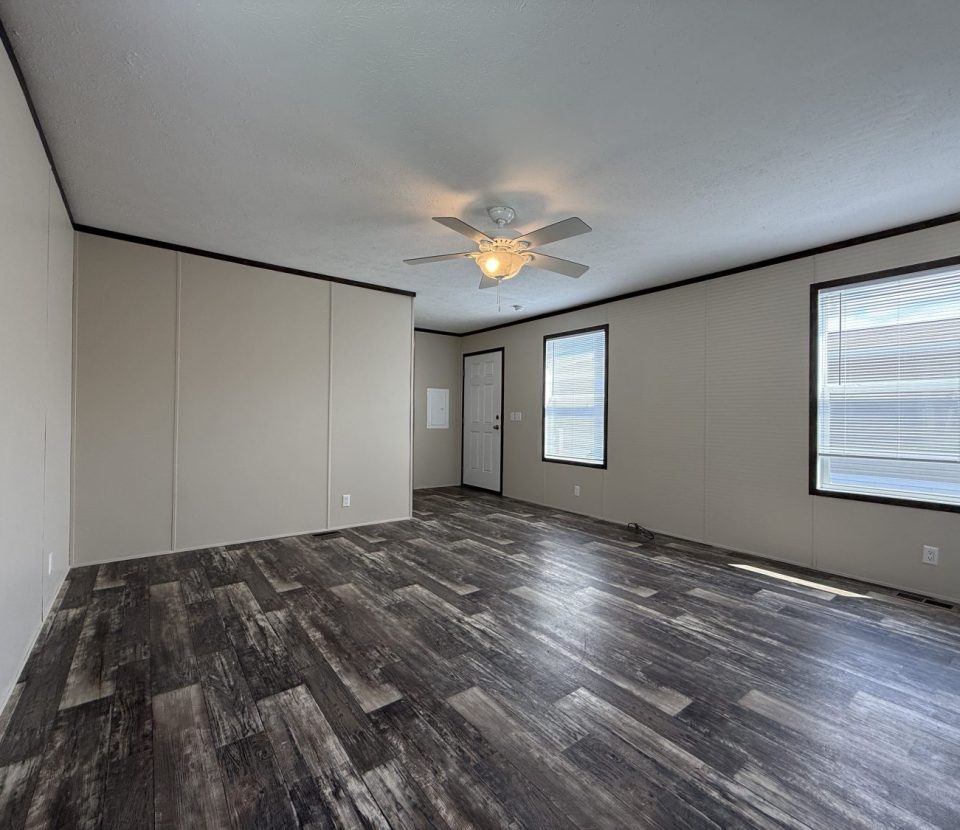 A spacious, empty room with light beige walls, dark trim, wood-look flooring, two large windows, a white ceiling fan with lights, and a closed door. Natural light fills the room.