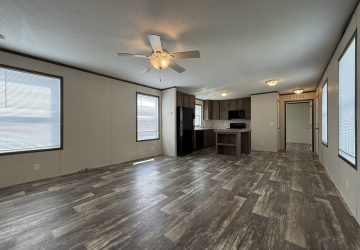 Spacious open-plan living area with wood-look flooring, ceiling fan, large windows, and a modern kitchen with dark cabinets and white countertops. Bright natural light fills the room.