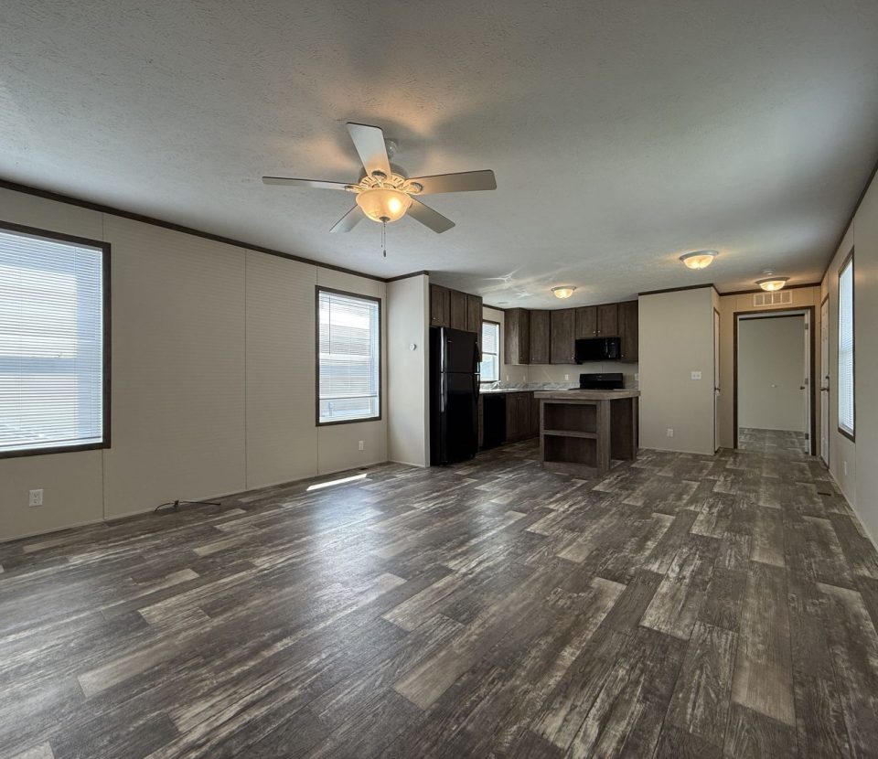 Spacious open-plan living area with wood-look flooring, ceiling fan, large windows, and a modern kitchen with dark cabinets and white countertops. Bright natural light fills the room.