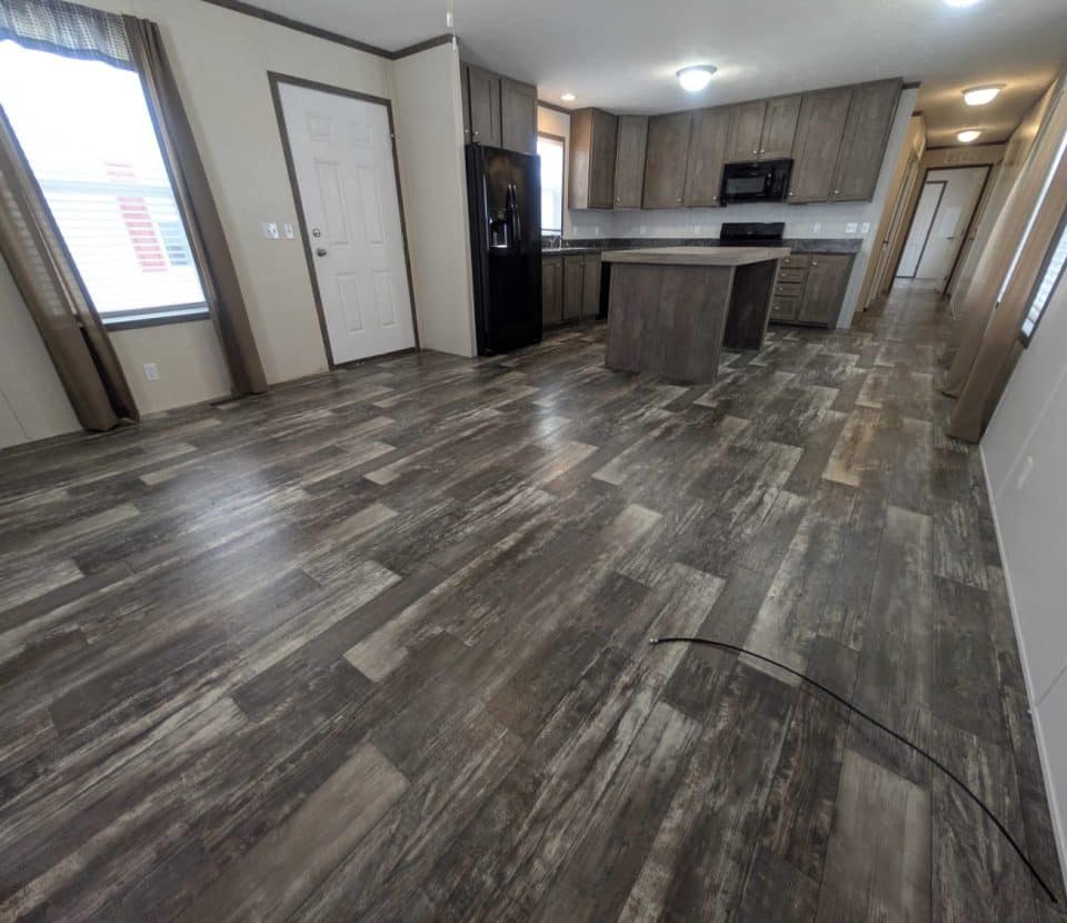 An empty, modern kitchen and living area with wood-look flooring, dark cabinets, a center island, and stainless steel appliances. Large windows and neutral walls add brightness to the open space.