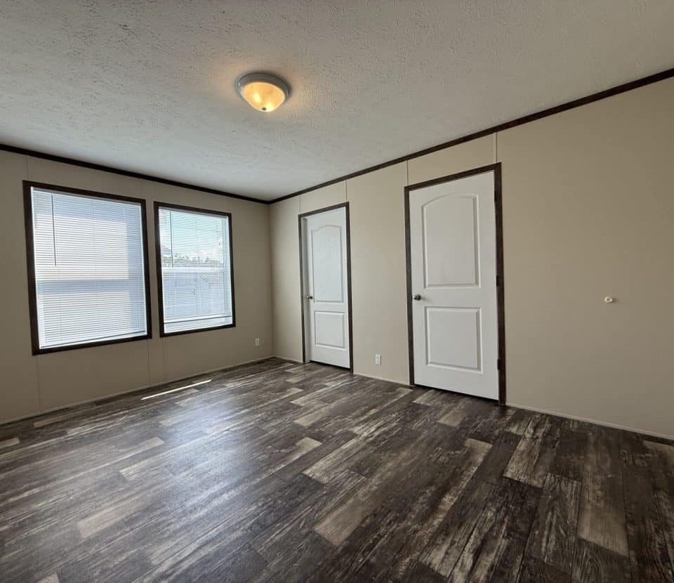 An empty room with beige walls, two white doors, two large windows with blinds, a ceiling light, dark trim, and wood-patterned flooring. Natural light fills the space.