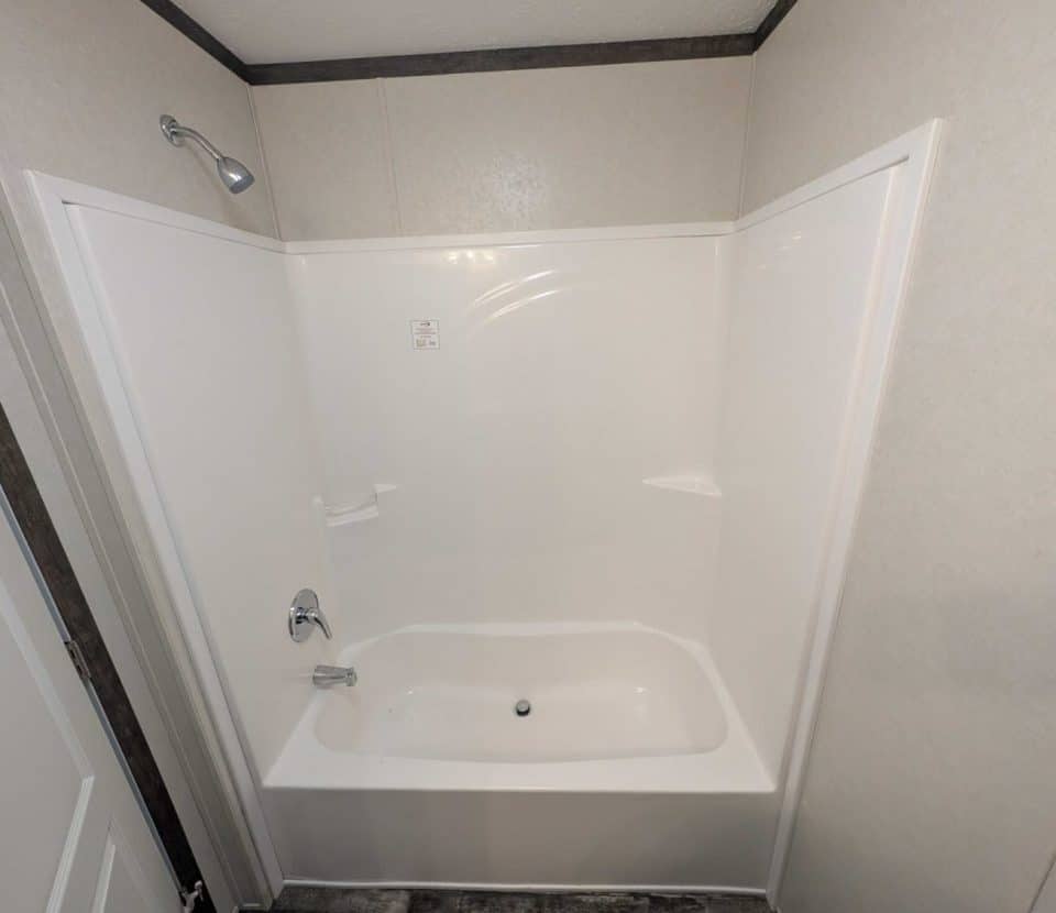 White bathtub and shower combo with a showerhead, faucet, and a small built-in soap holder, set against plain light-colored walls and a partial view of a door on the left side.