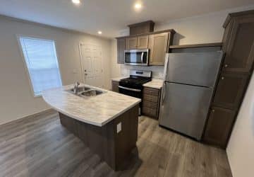 Modern kitchen with wood cabinets, stainless steel appliances, a marble-topped island with a sink, light wood flooring, and recessed lighting. A window and a white door are visible in the background.