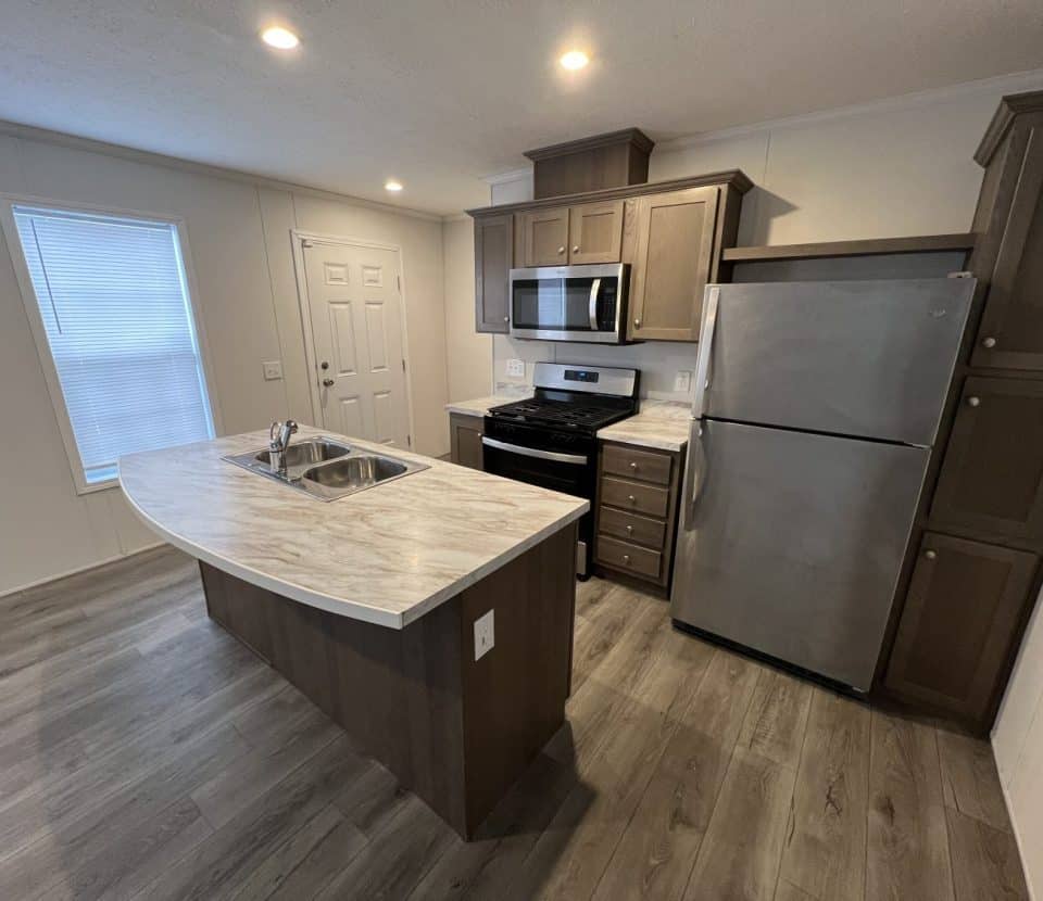 Modern kitchen with wood cabinets, stainless steel appliances, a marble-topped island with a sink, light wood flooring, and recessed lighting. A window and a white door are visible in the background.