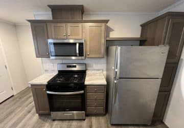 A compact kitchen with a gas stove, oven, microwave, refrigerator, and brown cabinets. The appliances are stainless steel, and the floor is wood-patterned laminate.