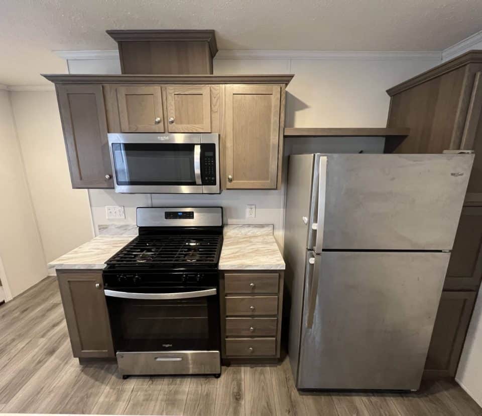 A compact kitchen with a gas stove, oven, microwave, refrigerator, and brown cabinets. The appliances are stainless steel, and the floor is wood-patterned laminate.