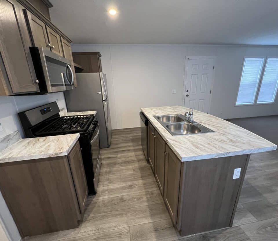 Modern kitchen with wood cabinets, marble-look countertops, a stainless steel stove and microwave, double sink on an island, and light wood flooring. Door and windows visible in the background.
