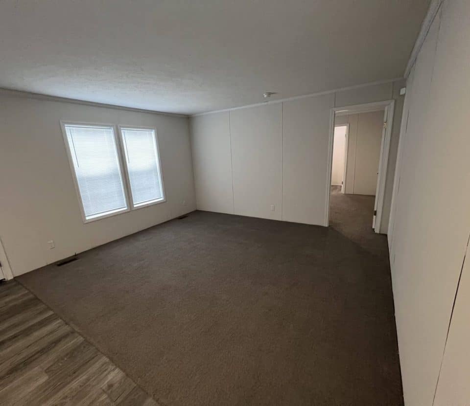 Empty room with beige carpet, white walls, two windows on the left, and open doorways leading to other rooms. The floor near the entrance is wood-style vinyl. The room is well-lit by natural light.