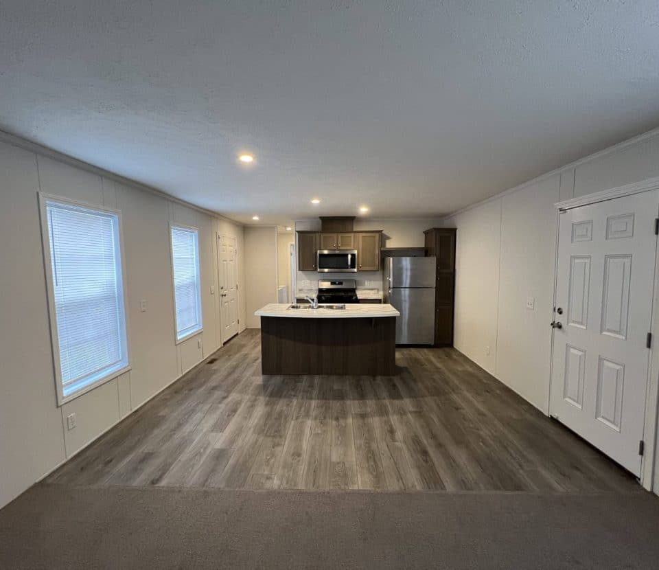 Modern kitchen with stainless steel appliances, white walls, and wood cabinets. An island is centered under recessed lighting. Large windows line the left wall, and a door is on the right. Floors are laminate and carpet.