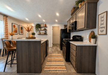 Modern kitchen with dark wood cabinets, marble countertops, black appliances, a patterned rug, decorative plants, and framed artwork on the walls. A dining area with brown chairs is visible to the left.