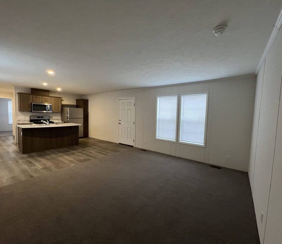 Modern open-plan living space with a kitchen on the left, featuring wood cabinets and stainless steel appliances, and a carpeted living area with large windows and white walls on the right.