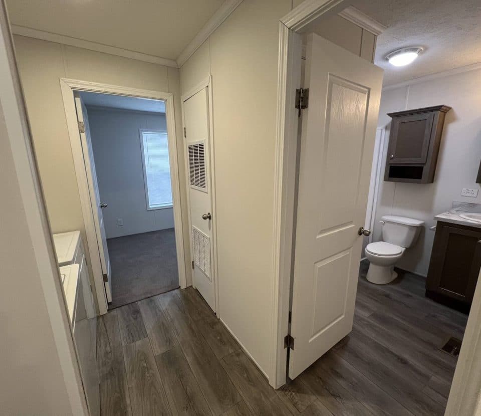 Hallway with wood flooring, a bathroom with a white toilet and sink on the right, a doorway leading to a carpeted room in the back, and laundry area on the left. Light walls and white doors throughout.