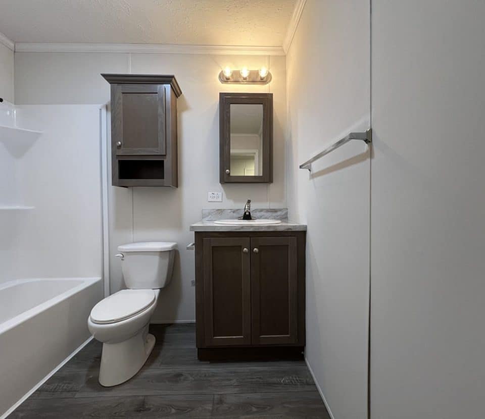 Bathroom with a toilet, bathtub, sink, wooden cabinets, wall mirror, towel rack, and three-bulb vanity light on white walls and dark wood-look flooring.