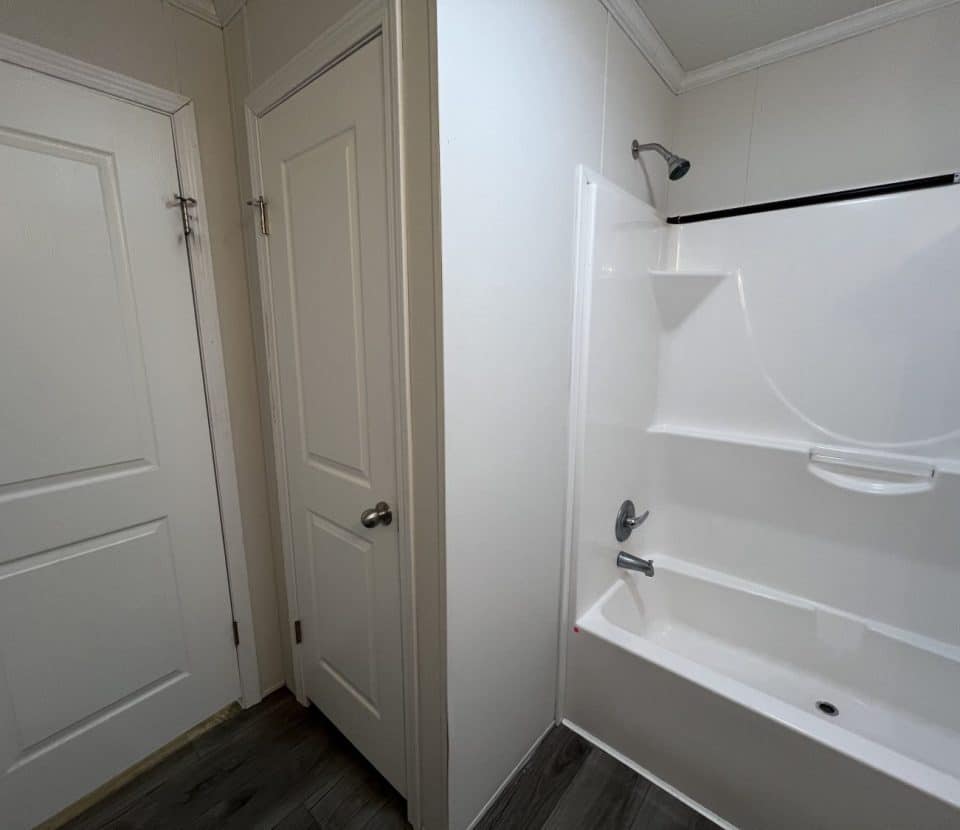 A bathroom with two white doors, a white bathtub-shower combo with shelves, a showerhead, faucet, and gray wood-style flooring. The walls are light-colored.