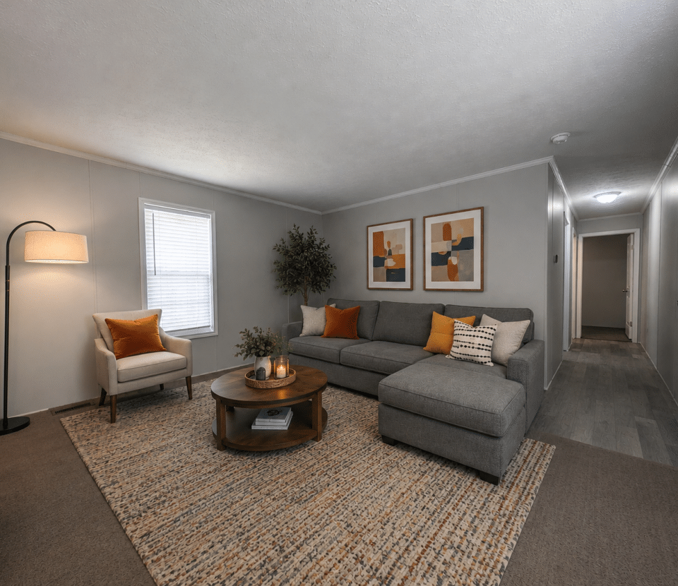 A cozy living room with a gray sectional sofa, armchair, round wooden coffee table, floor lamp, two abstract art pieces on a gray wall, and a hallway leading to the back. Neutral colors and warm accents decorate the space.