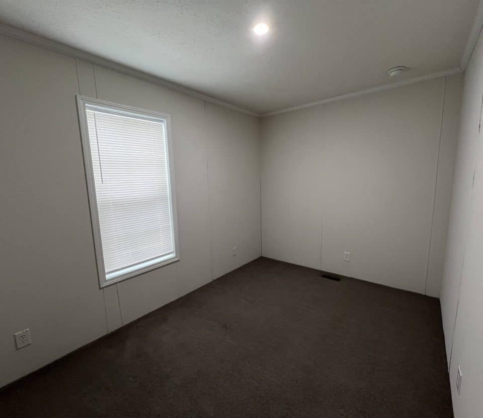 Empty room with white walls, a single window with blinds, dark carpeted floor, a ceiling light, and electrical outlets on the walls. The space appears clean and simple.