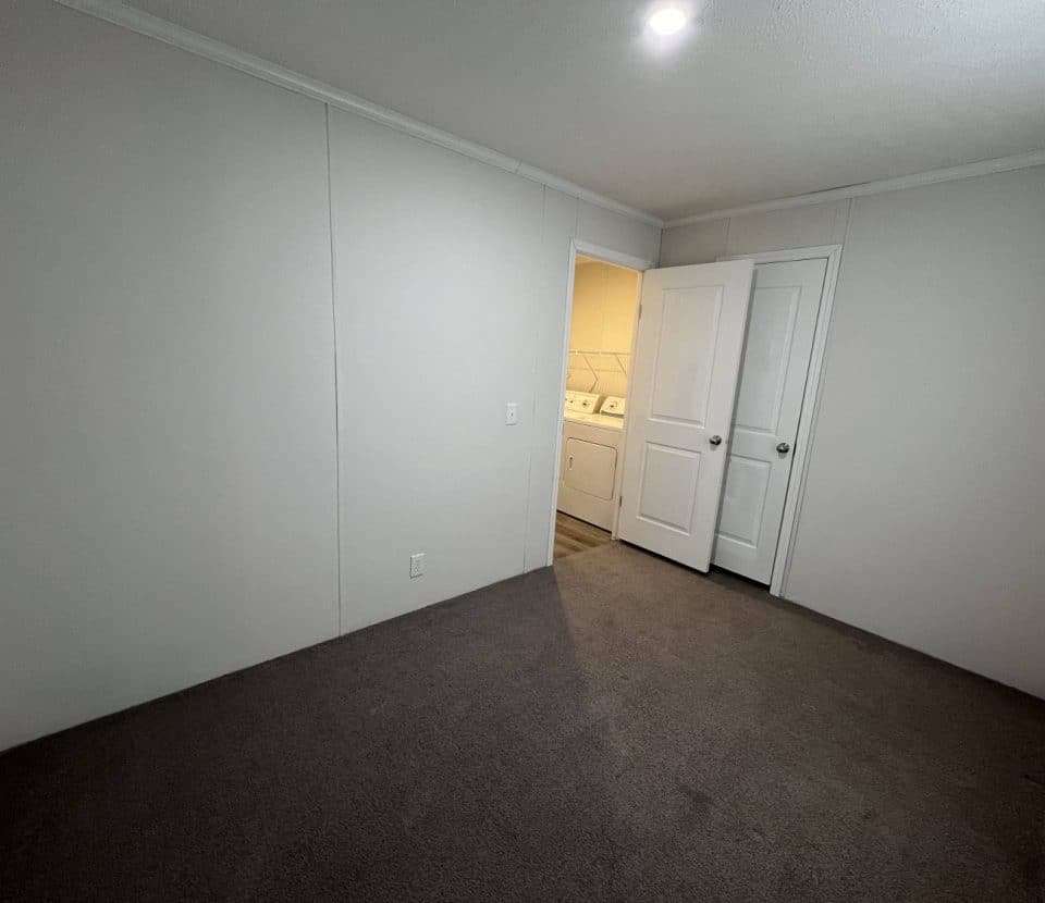Empty room with gray carpet, white walls, and a ceiling light. There are double doors open to reveal a laundry area with a washer and dryer and bright lighting.